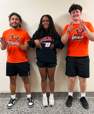  Three smiling young people indoors making a hand gesture with both hands, forming the letter "U."